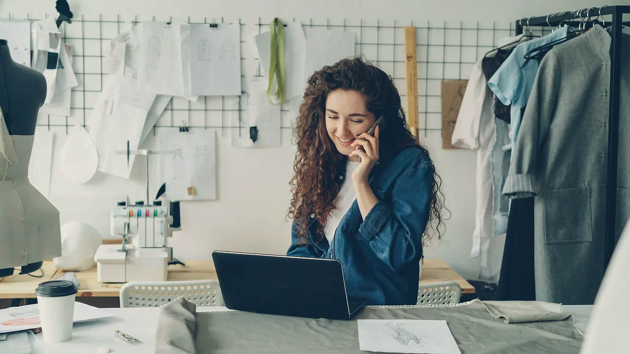 A woman in a sewing studio smiling on a phone call at her laptop, with sketches pinned to the wall behind her